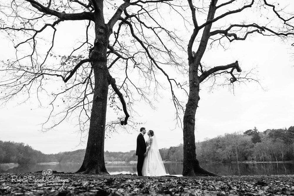 Bride and groom standing together between two large trees on the shore of a lake