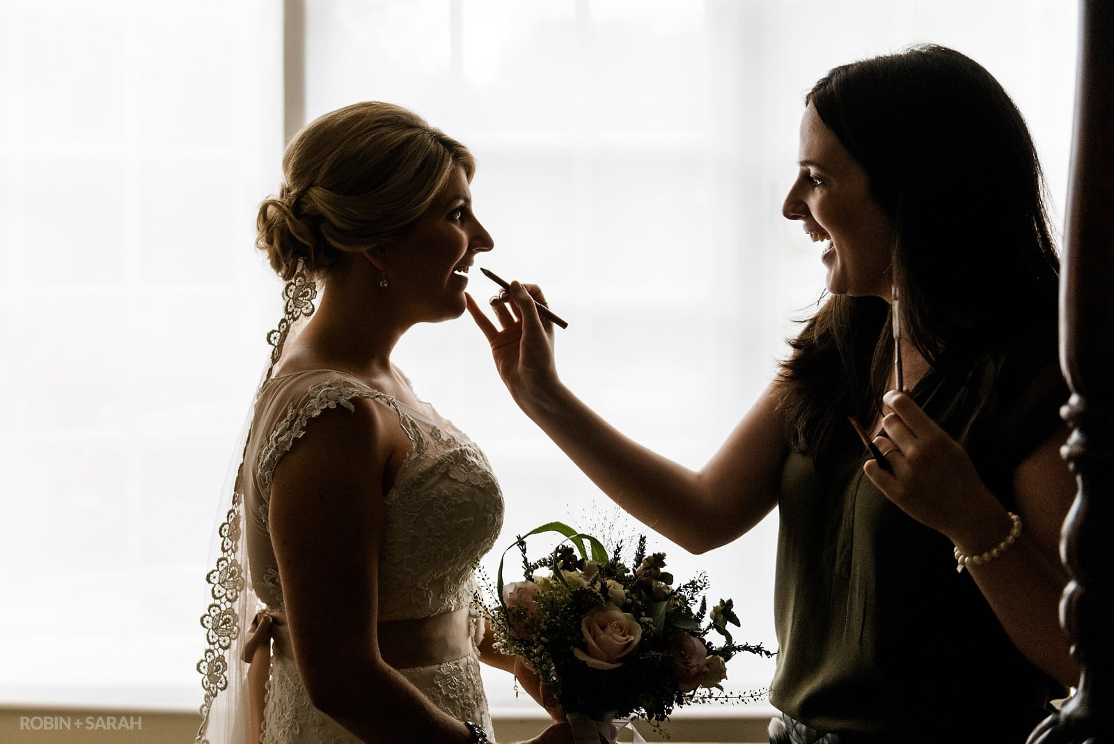 Bride silhouetted in window light as she has makeup applied