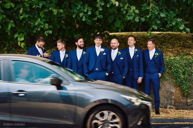 Groom and groomsmen laughing during group photo as car passes