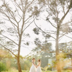 Two brides holding hands beneath two tall trees