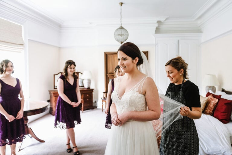 Bride has veil fitted in bedroom at Pendrell Hall as bridesmaids watch