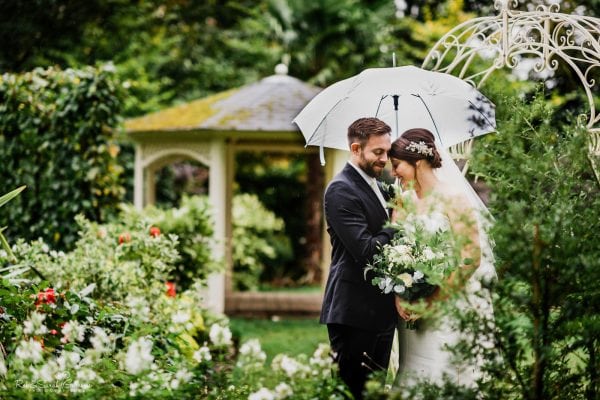 Bride and groom under white umbrella in gardens at Warwick House on rainy day
