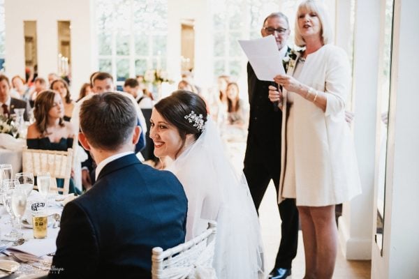 Bride smiles at groom as parents give wedding speech