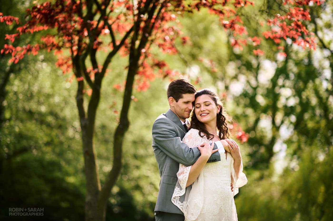 Groom holds bride as they share a quiet moment during garden wedding, with beautiful red tree in background