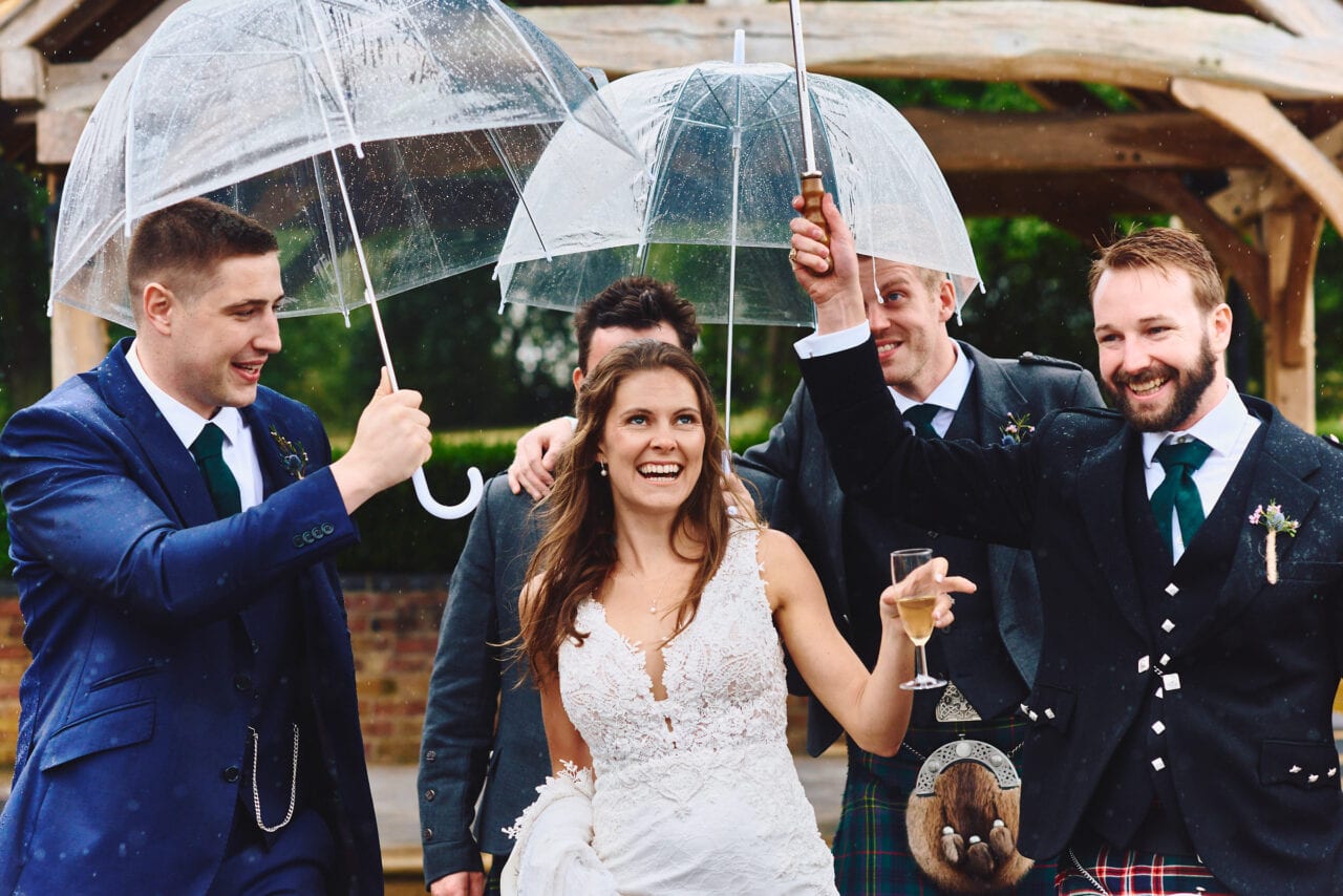 Bride laughing as friends hold umbrellas over her in the rain