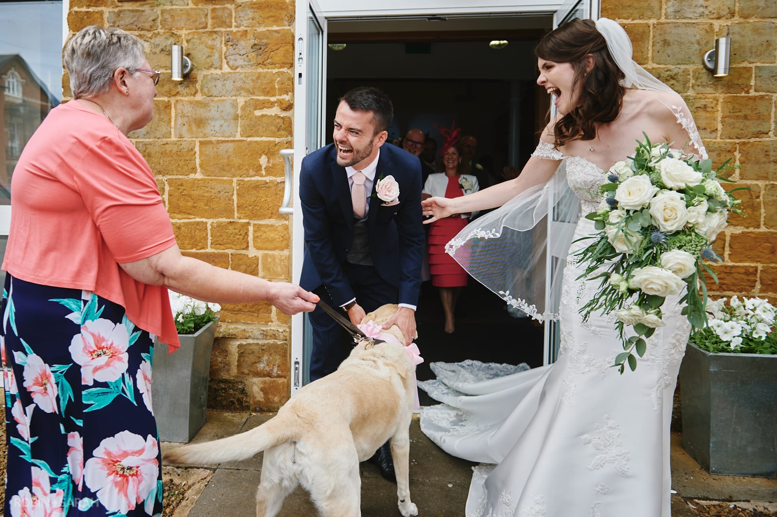 Bride and groom laughing as friend meets them with dog outside church wedding ceremony