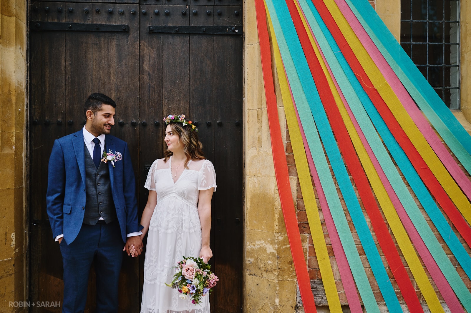 Bride and groom hold hands in doorway of old building with coloured ribbons
