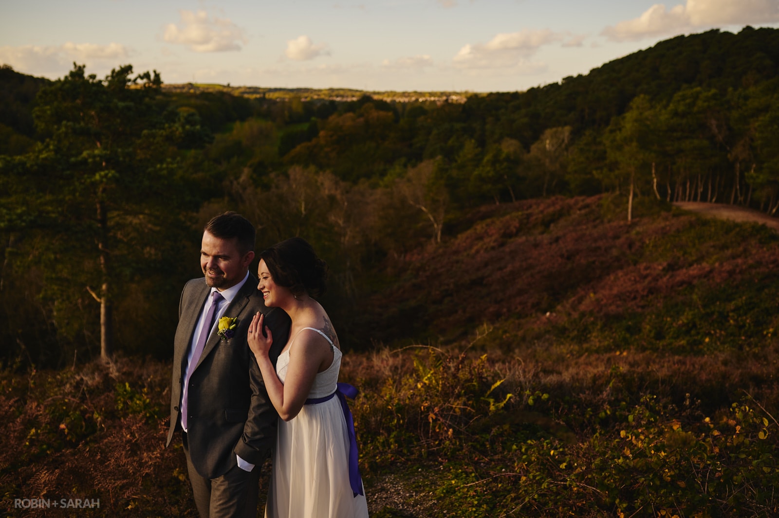 Bride and groom share a quiet moment standing together on top of a hill surrounded by lush woodlands in beautiful autumnal light