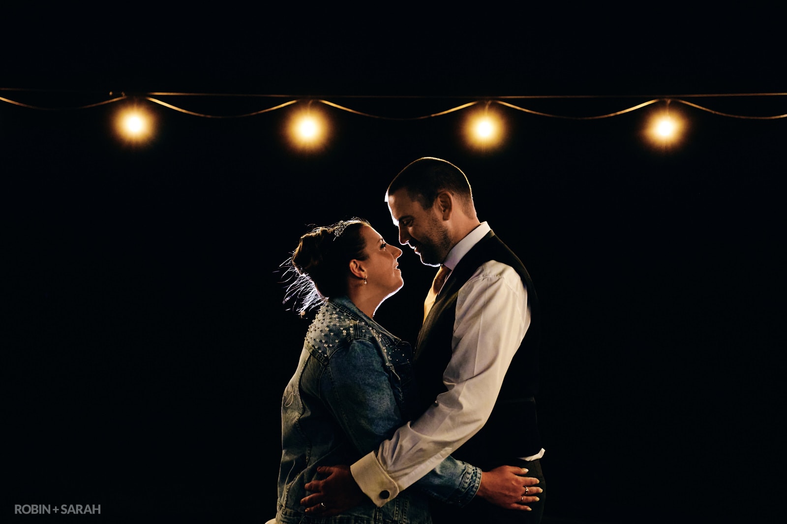 Bride and groom face each other under festoon light at The Barn in Dinthill