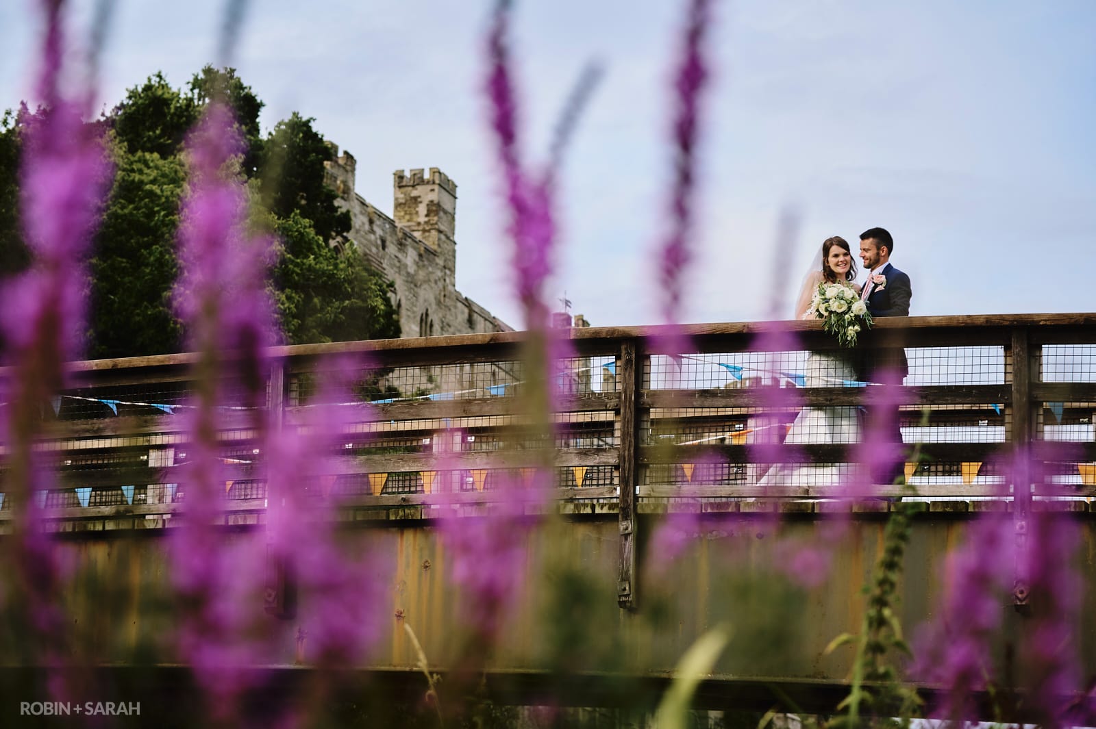 Bride and groom on bridge in grounds of Warwick Castle with purple flowers in foreground