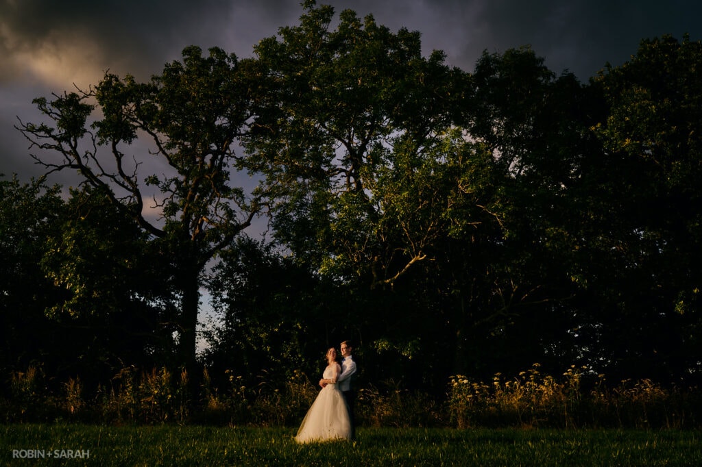 Bride and groom in fields at Wethele Manor, standing in front of tall trees with stormy sky