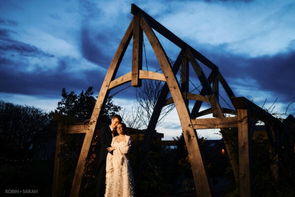 Bride and groom stand under wooden arch outside Cider Mill Barns at night
