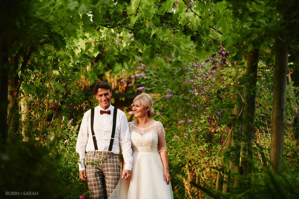 Bride and groom walk through pergola covered in plants and flowers at Oakfield Gardens