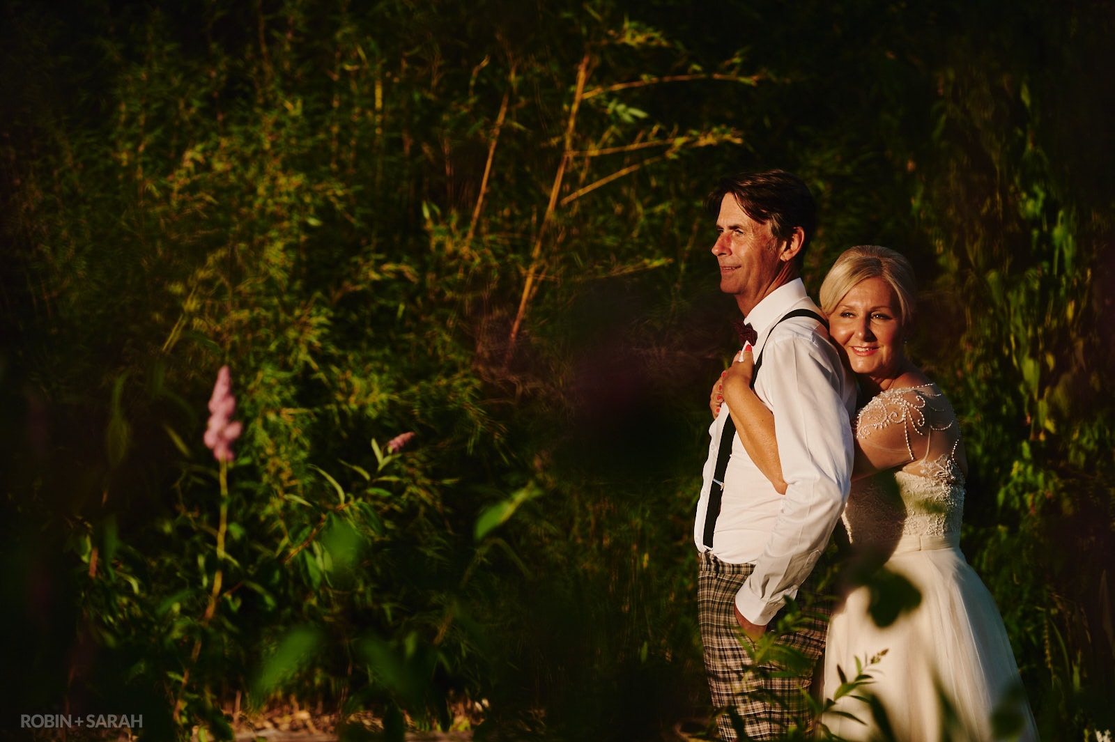 Bride hugs groom from behind as they stand in beautiful gardens with evening light