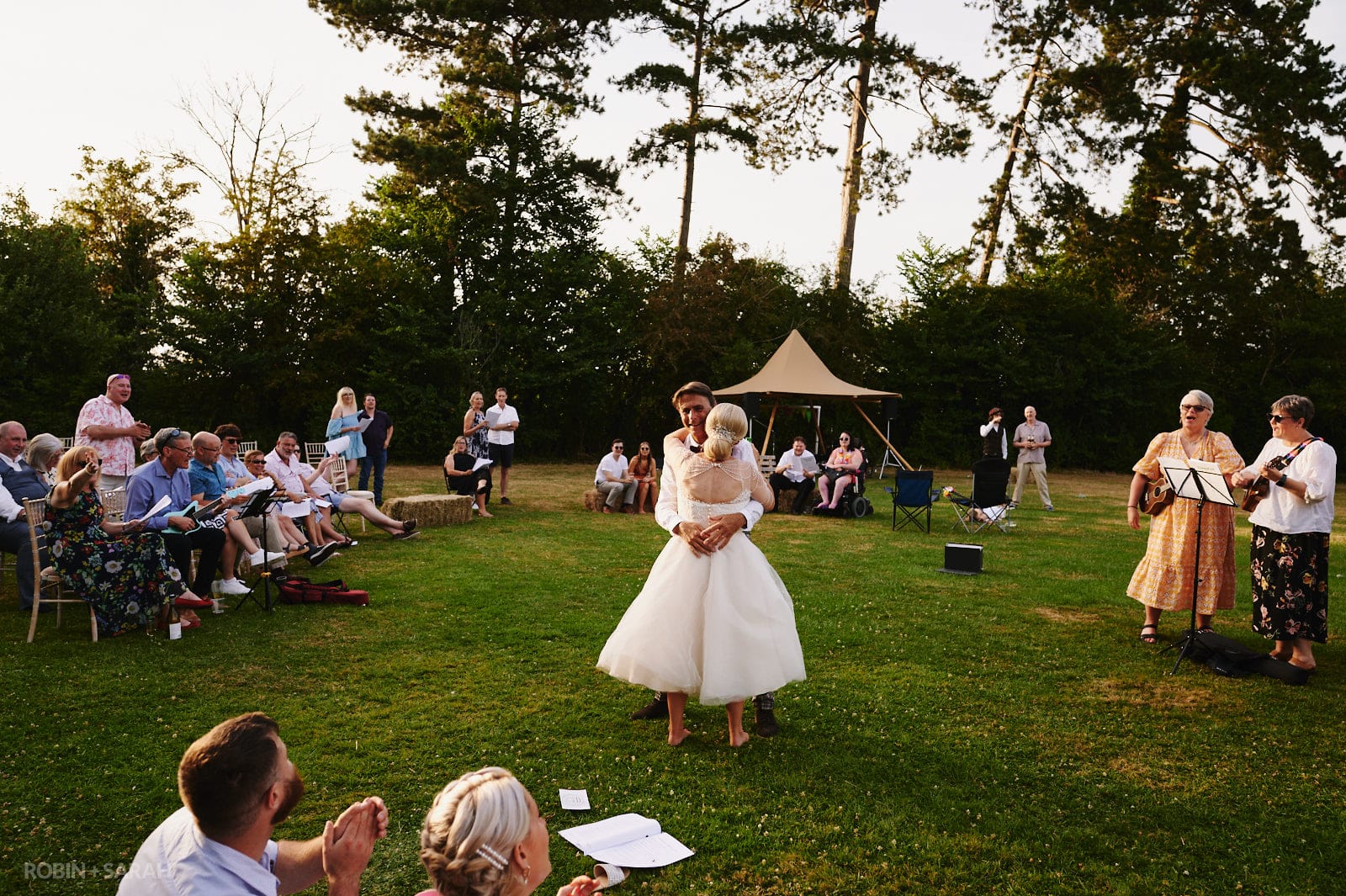 Bride and groom dance in field as wedding guests sing to live music
