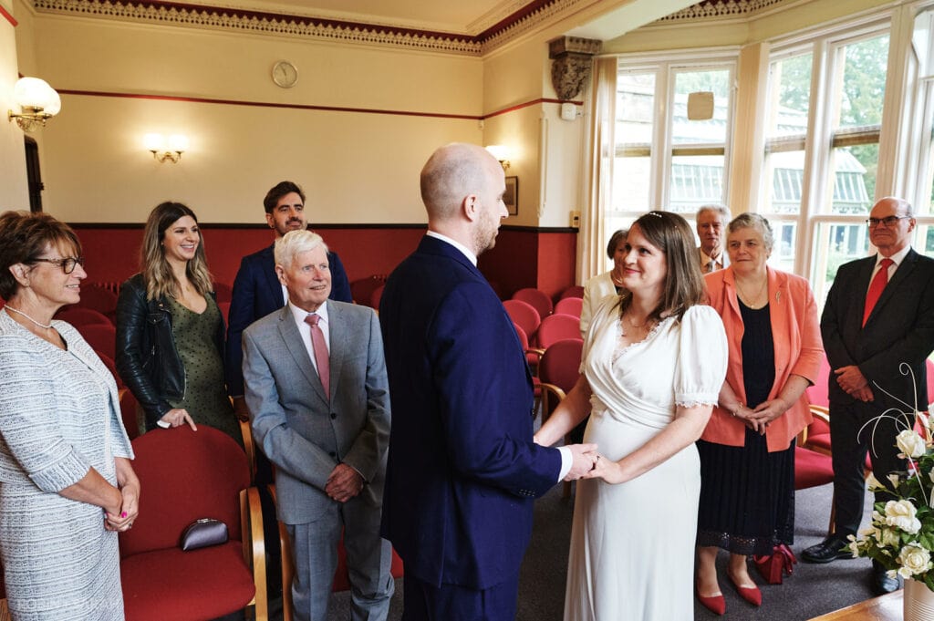 Bride and groom hold hands and exchange vows as guests watch in wedding ceremony in The Elgar Room at Malvern Registry Office