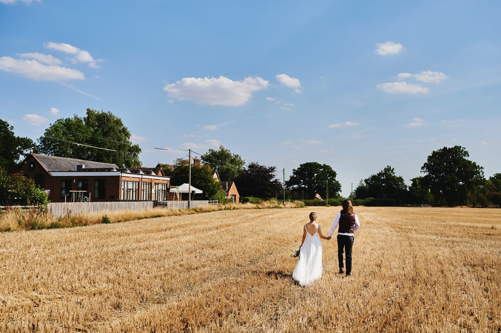 Bride and groom walk hand in hand through field at Bentley Village Hall