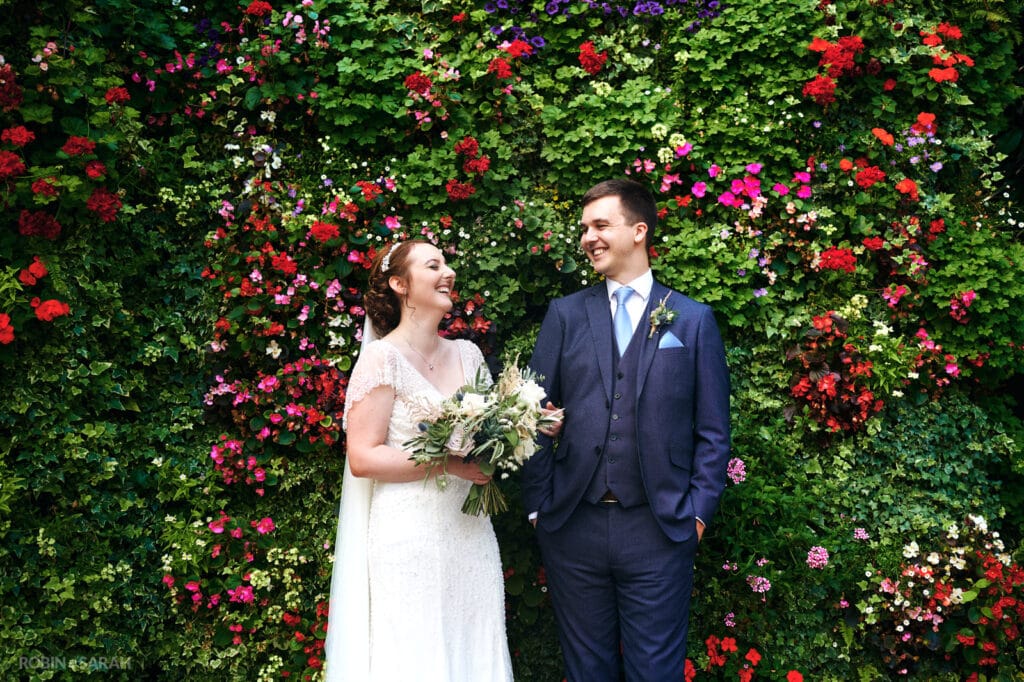 Bride and groom laughing together in front of flower wall at Moxull Hall