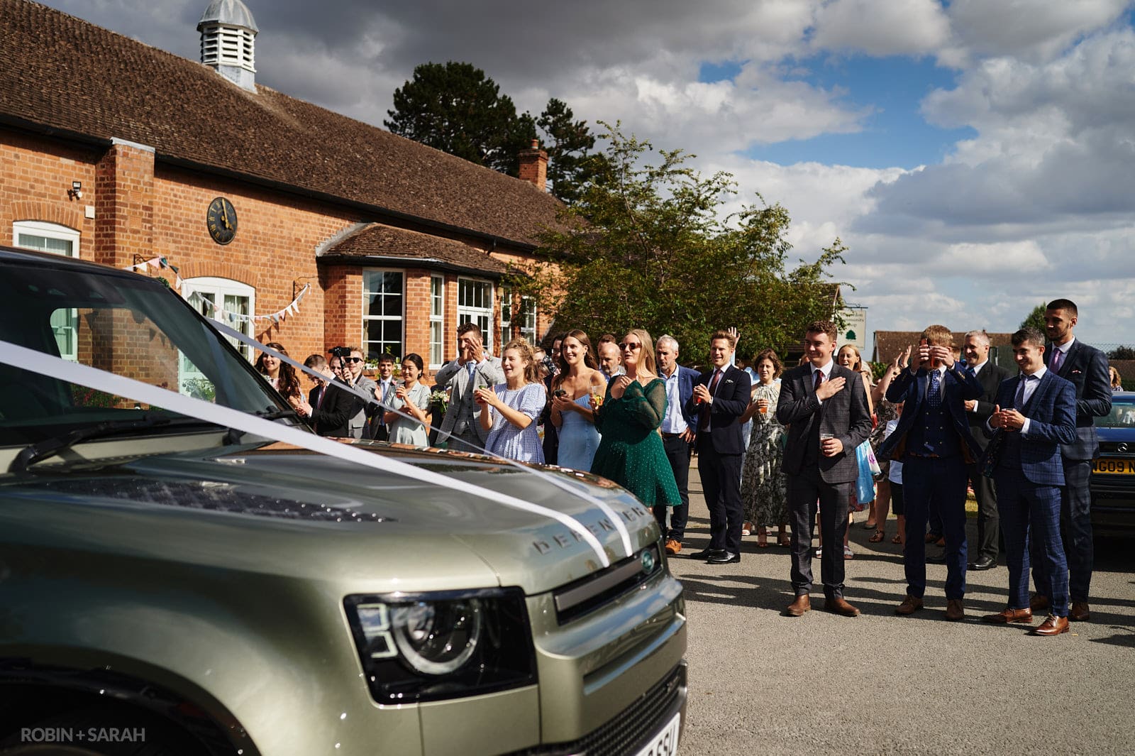 Wedding guests clap as bride and groom arrive in car at village hall