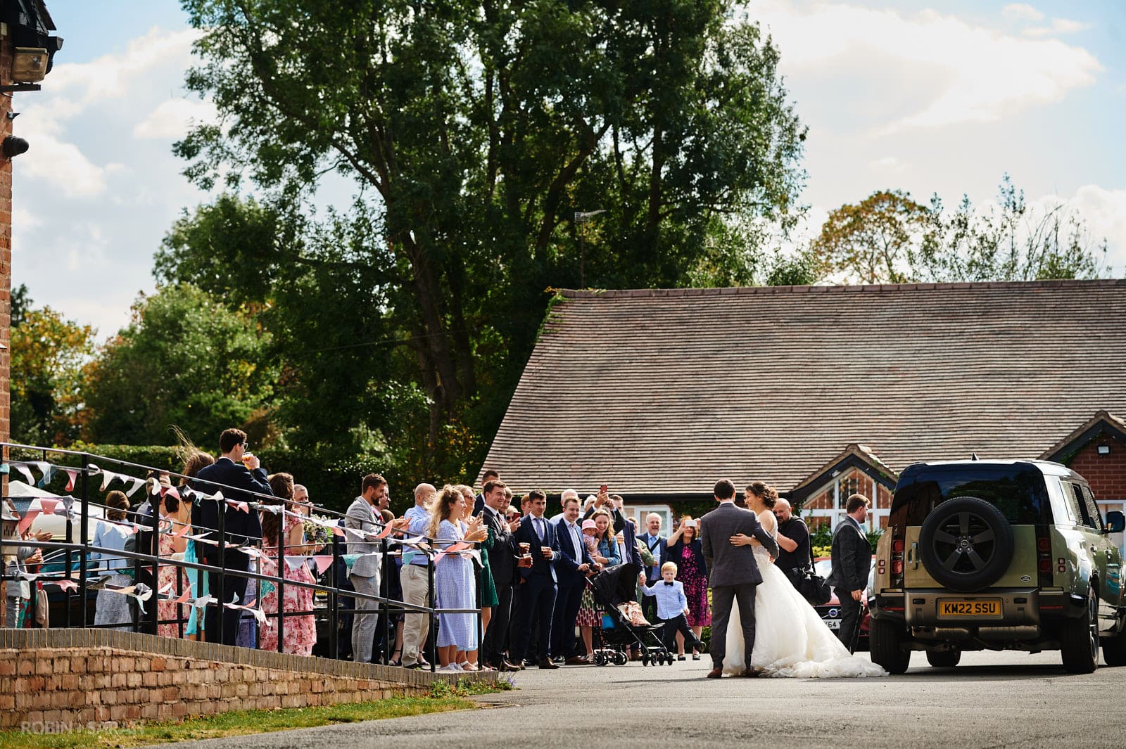 Bride and groom arrive at village hall for wedding reception as guests clap and cheer