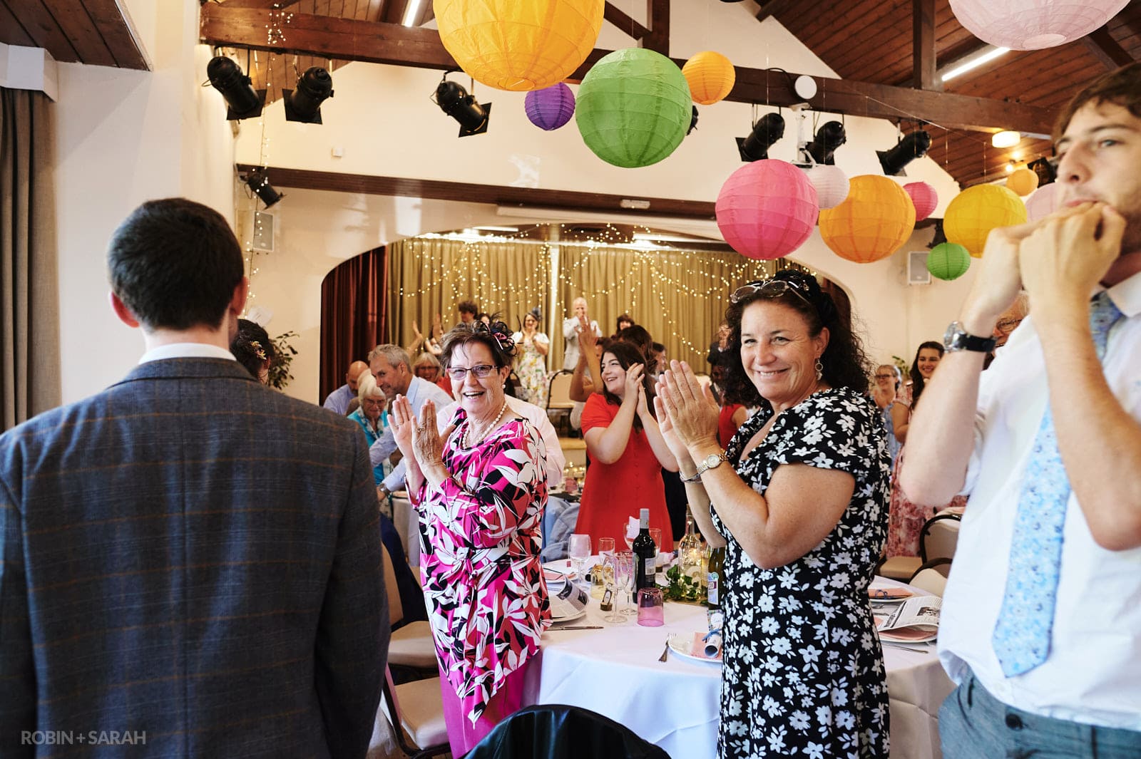 Wedding guests clap as bride and groom enter village hall for wedding reception