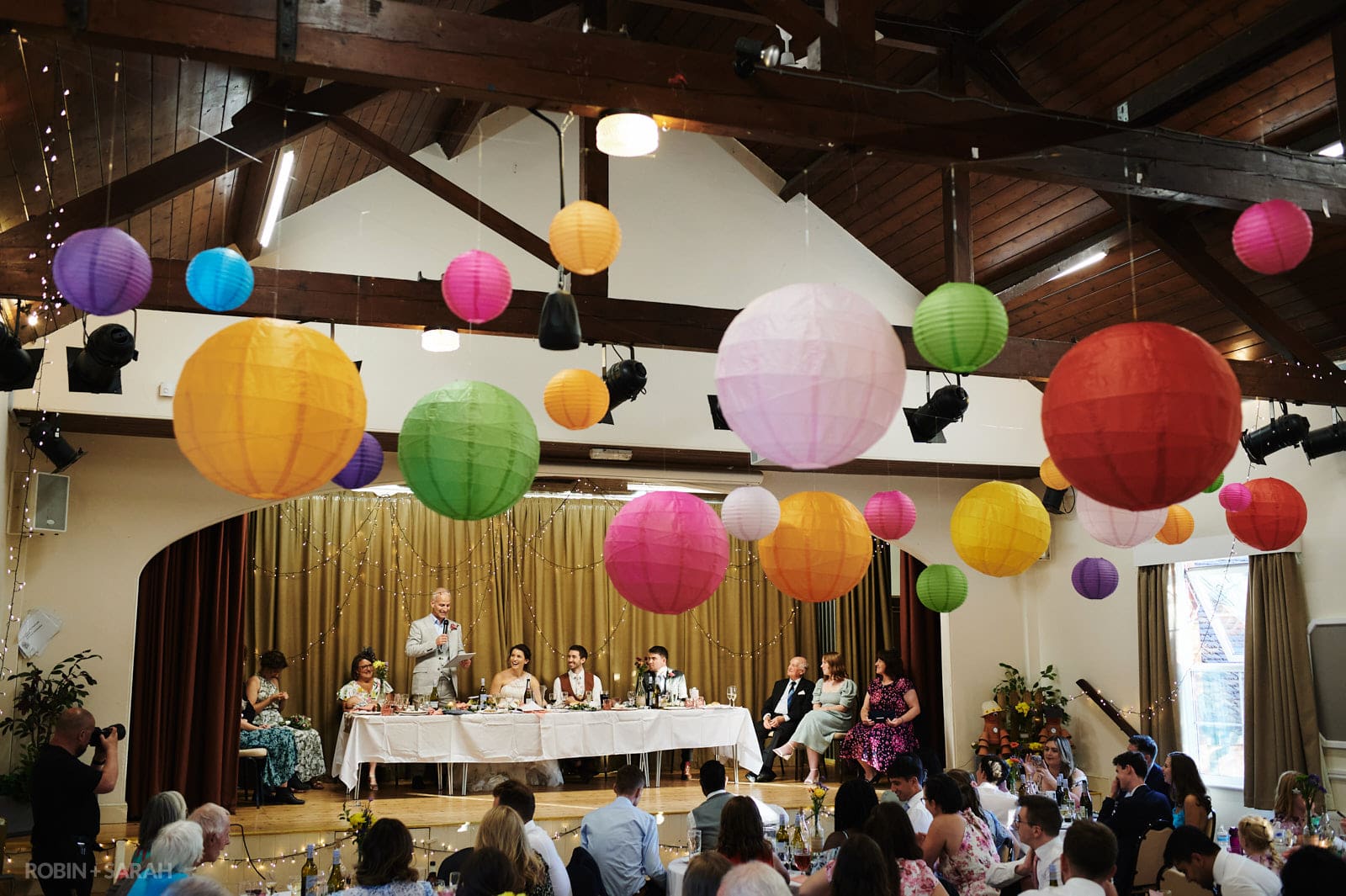 Wedding speeches in village hall with brightly coloured paper lanterns hanging from roof