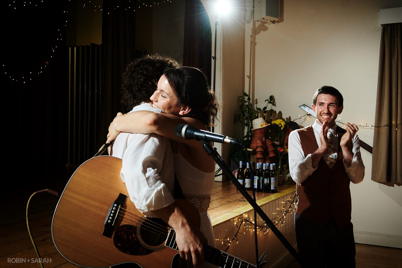 Bride hugs brother as he plays guitar and sings at her wedding in a village hall