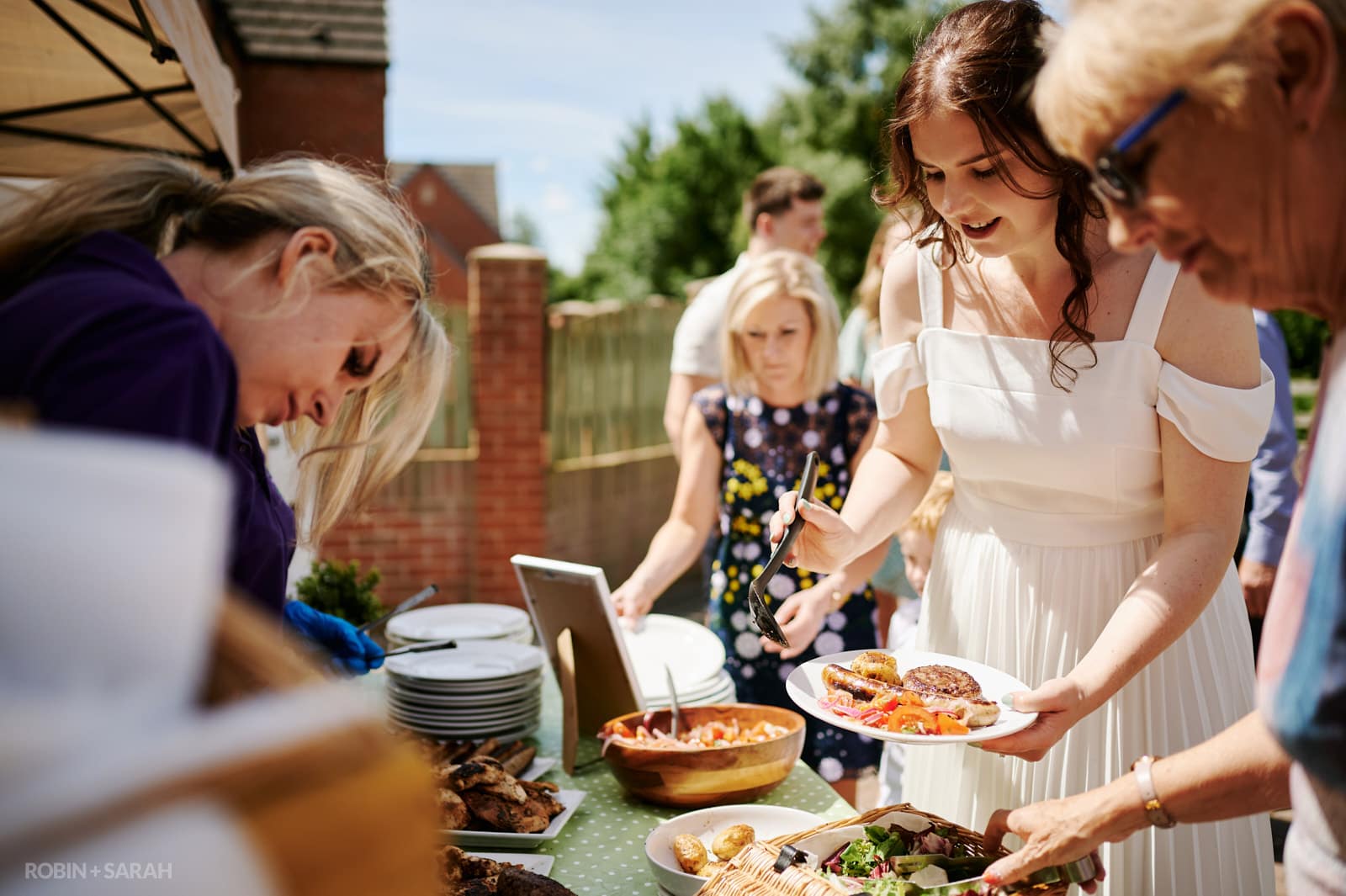 Bride and guests put freshly cooked food on plates at home wedding