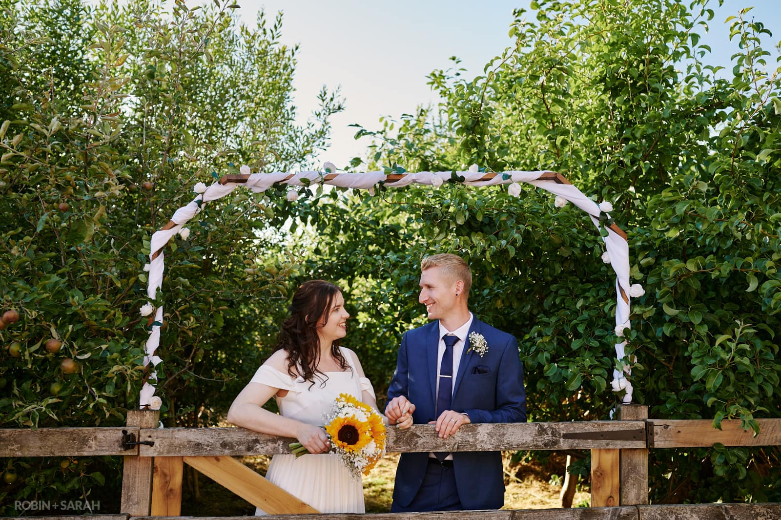 Bride and groom leaning on wooden fence and surrounded by bunting and greenery