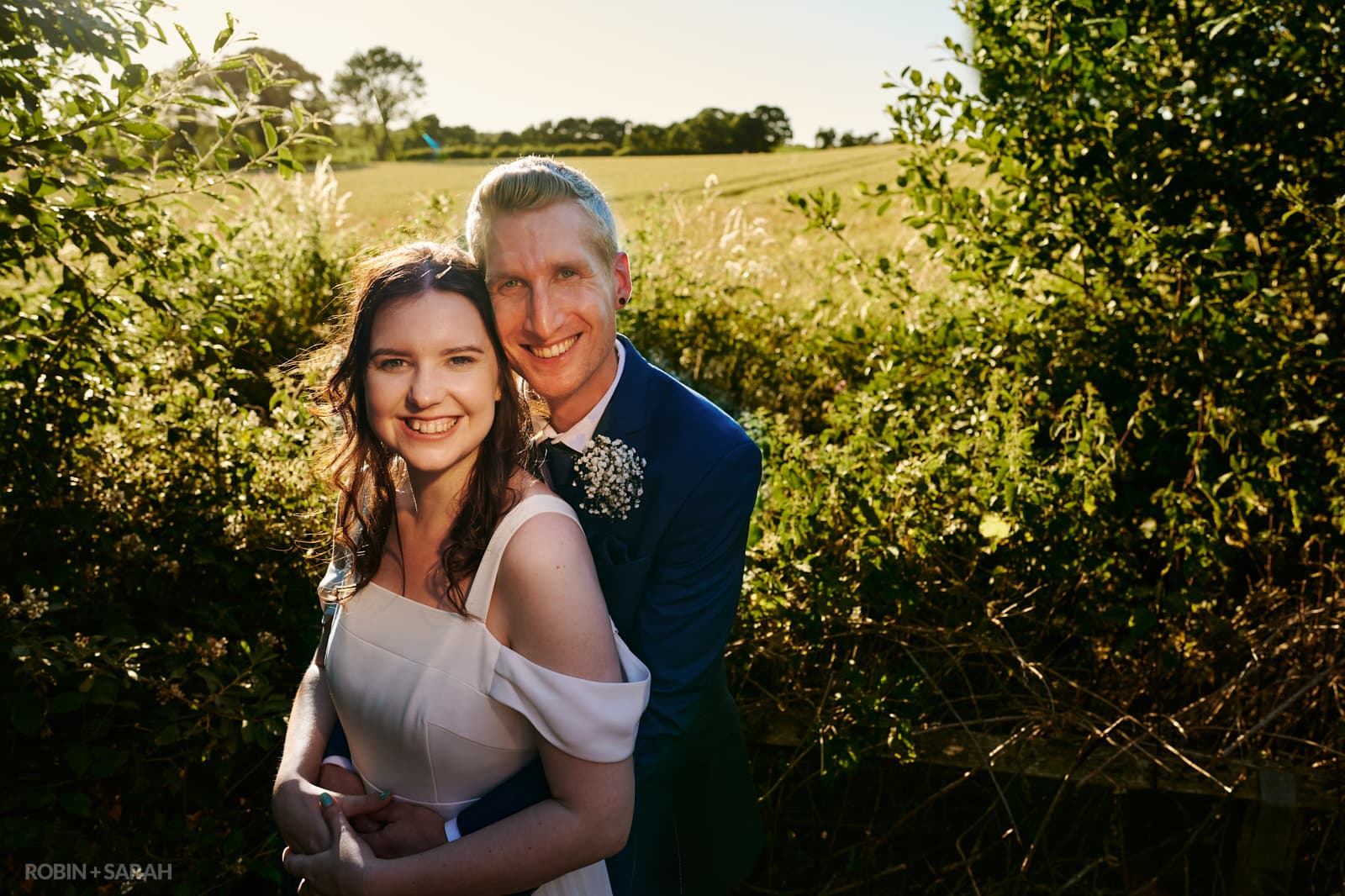 Bride and groom surrounded by trees and shrubs with crop field in background