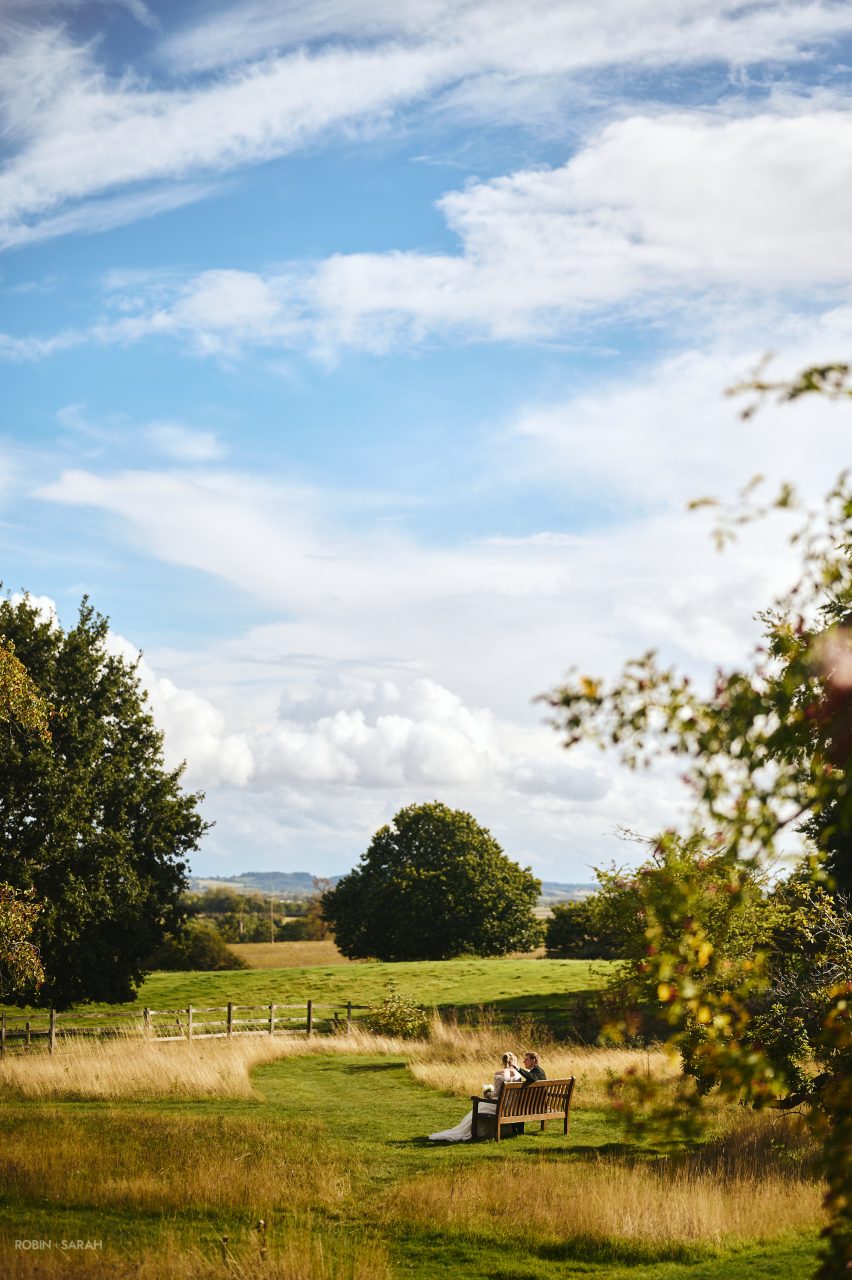 Bride and groom sitting on a bench amongst the long grass in grounds at Bourton Hall, with a blue sky and sunny conditions