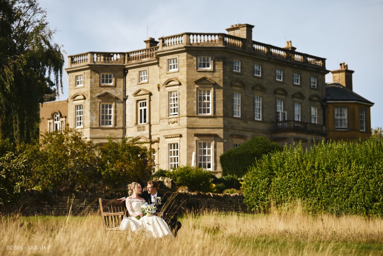 Newly married couple sit on bench in long grass, with Bourton Hall in the background