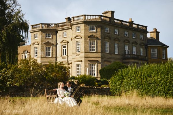 Newly married couple sit on bench in long grass, with Bourton Hall in the background