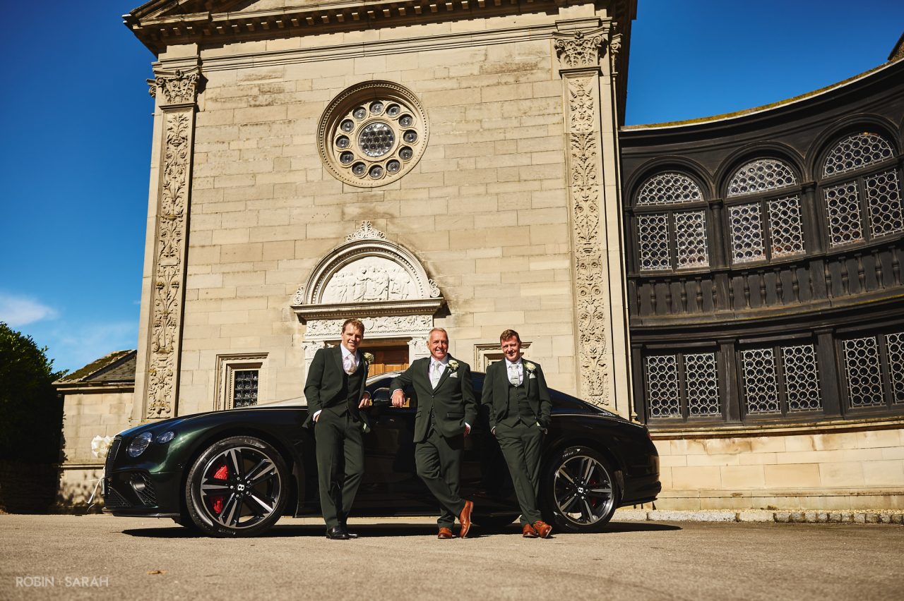 Groom, dad and best man posing for photo with Bentley wedding car, in front of the chapel at Bourton Hall