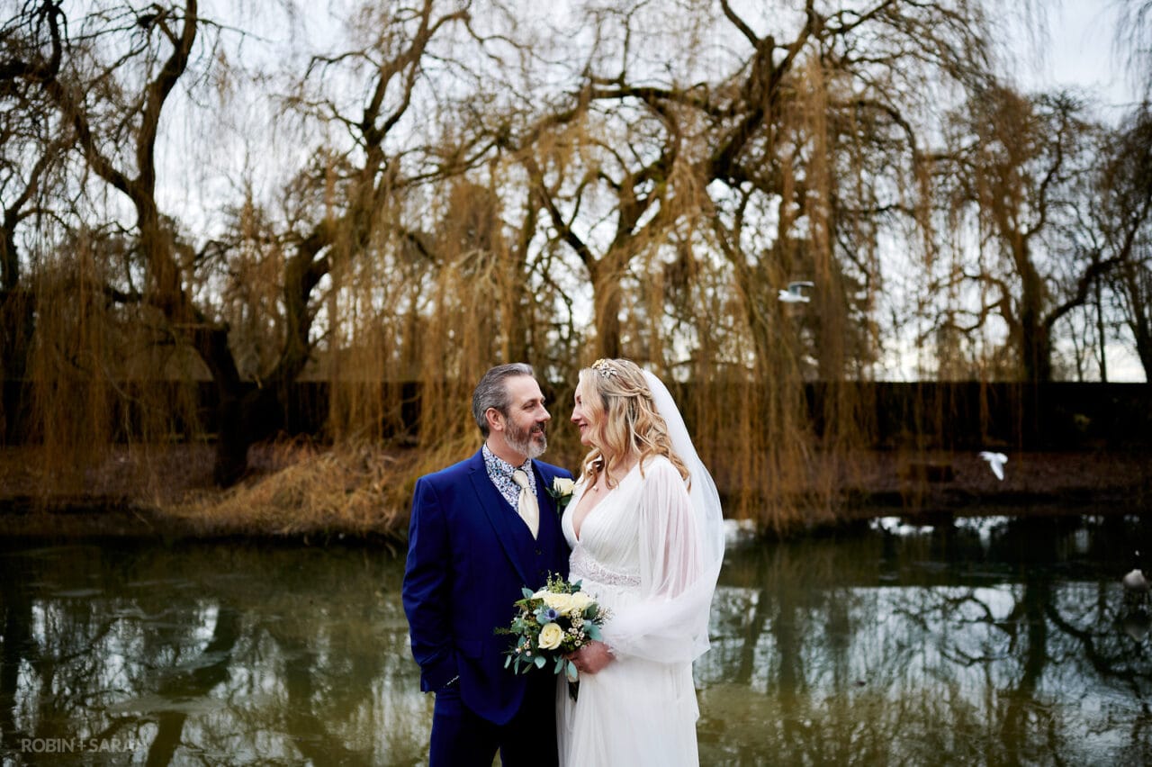 Newly married couple standing in front of lake at Coombe Abbey with weeping willow trees in background