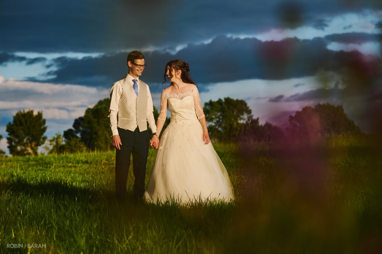 Bride and groom walk hand in hand through the fields at Wethele Manor with a dramatic, brooding sky behind them
