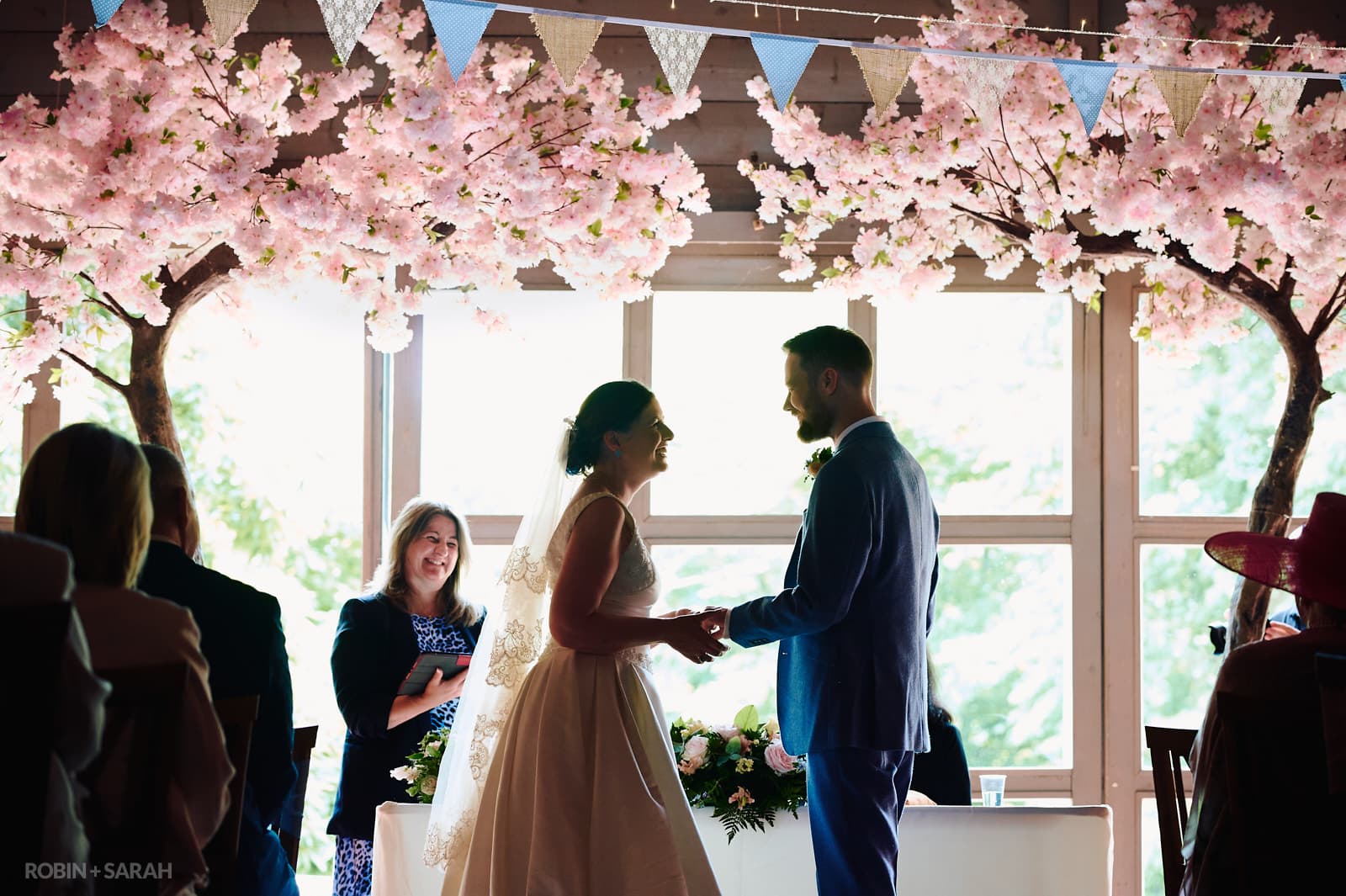 Bride and groom exchange wedding rings under pink decorative trees in Guesten Hall wedding ceremony.