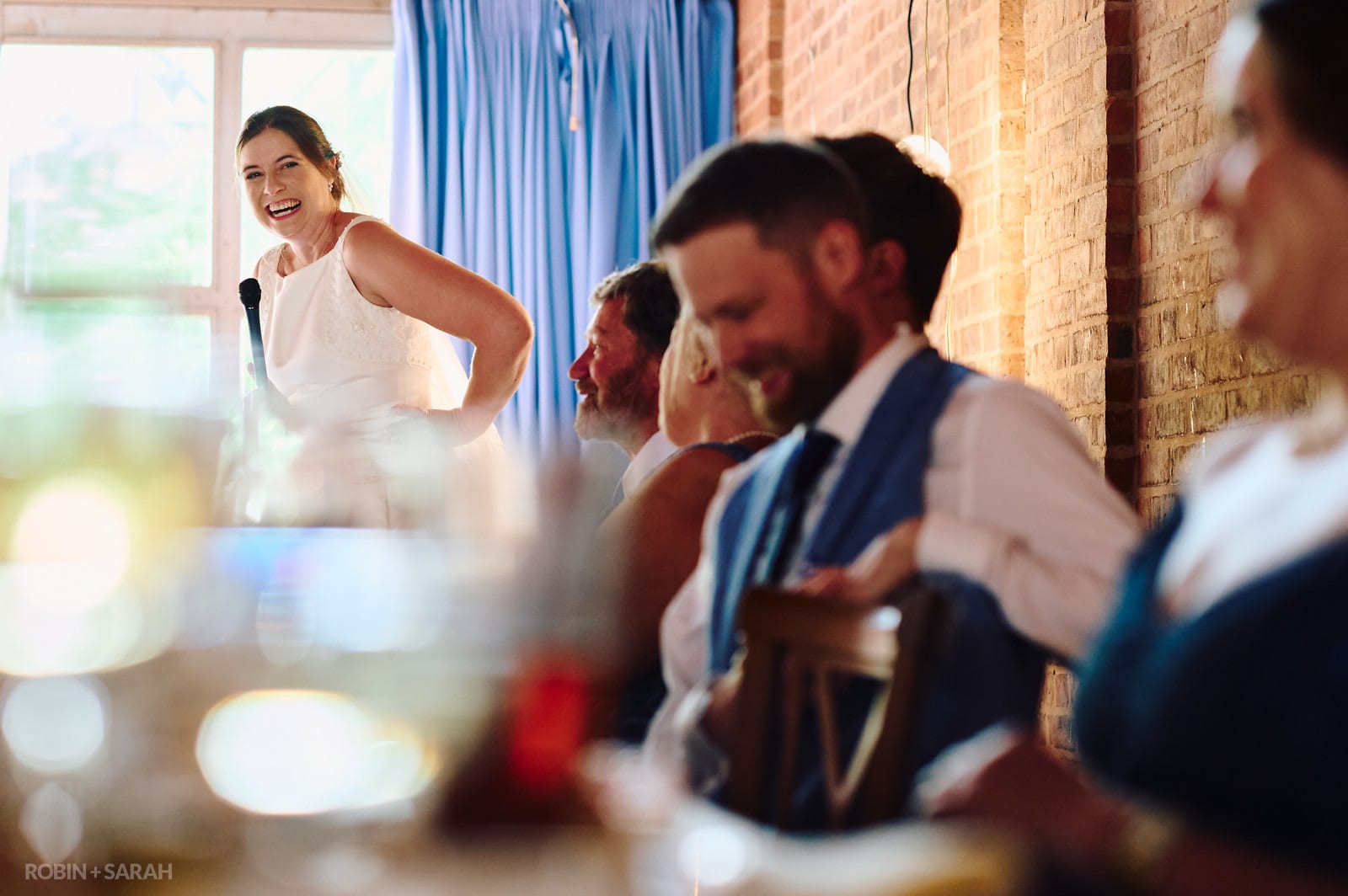 Bride gives wedding speech as groom and guests laugh