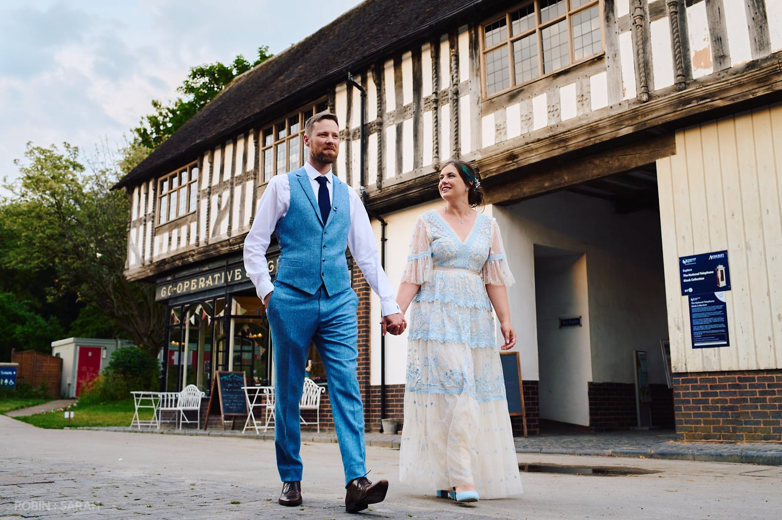 Bride in blue and white summery dress walks hand in hand with groom at Avoncroft Museum
