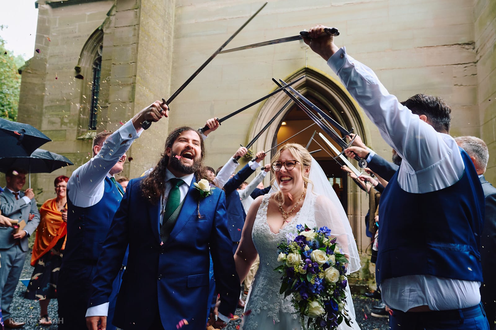 Bride and groom walk under sword arch at St Andrews church Ombersley