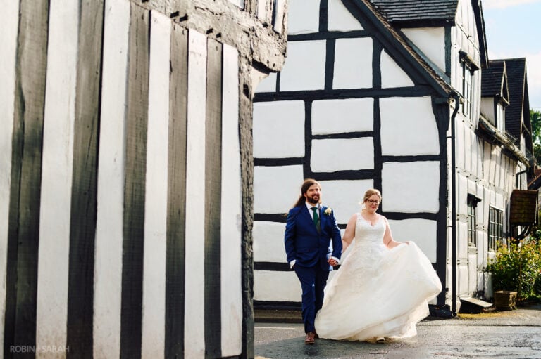 Bride and groom walk between old black and white half-timber buildings in Ombersley