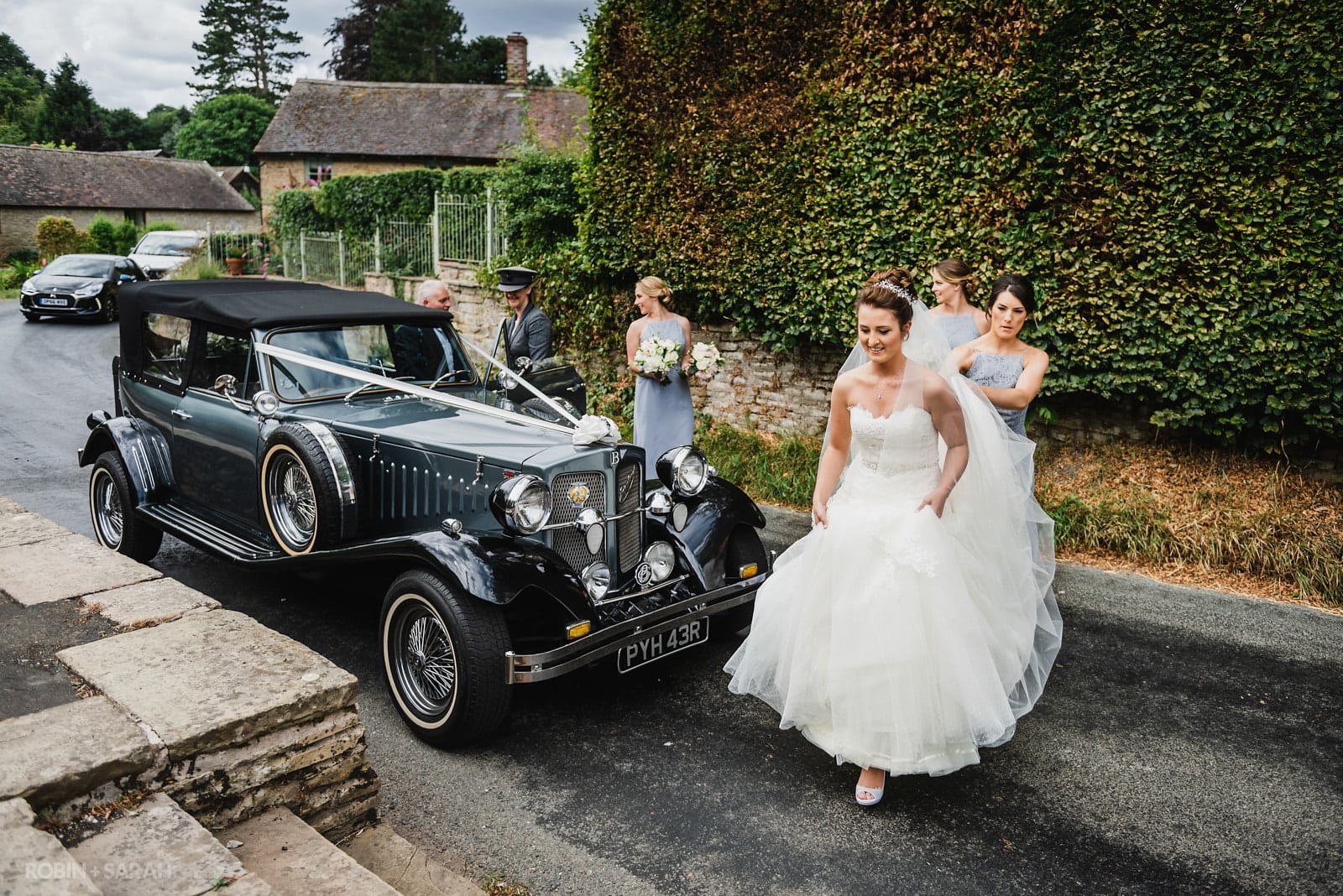Bride walks from wedding car to church as bridesmaids help her with dress and veil