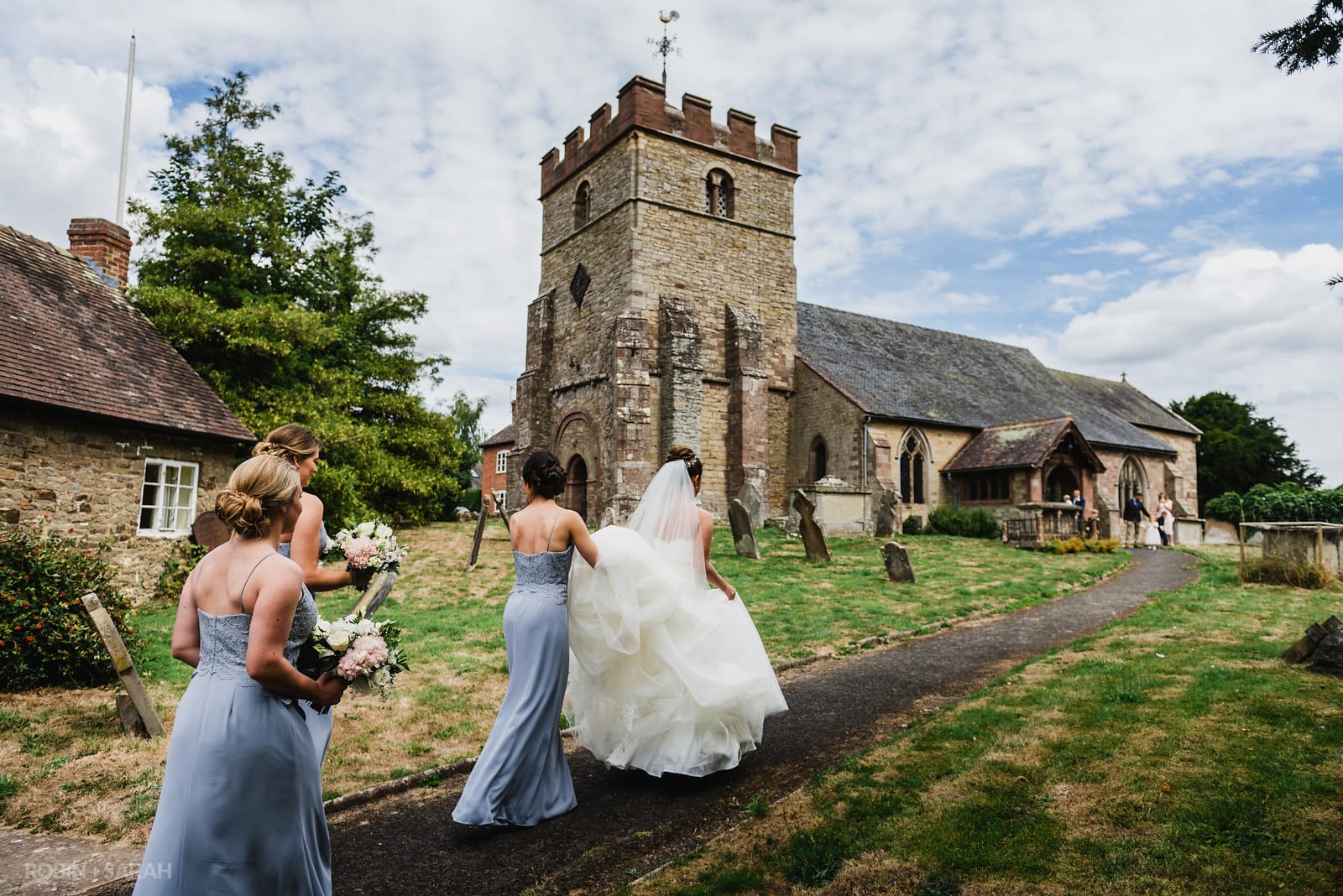 Bride walks up path to St Peter's Church in Diddlebury as bridesmaids help with dress