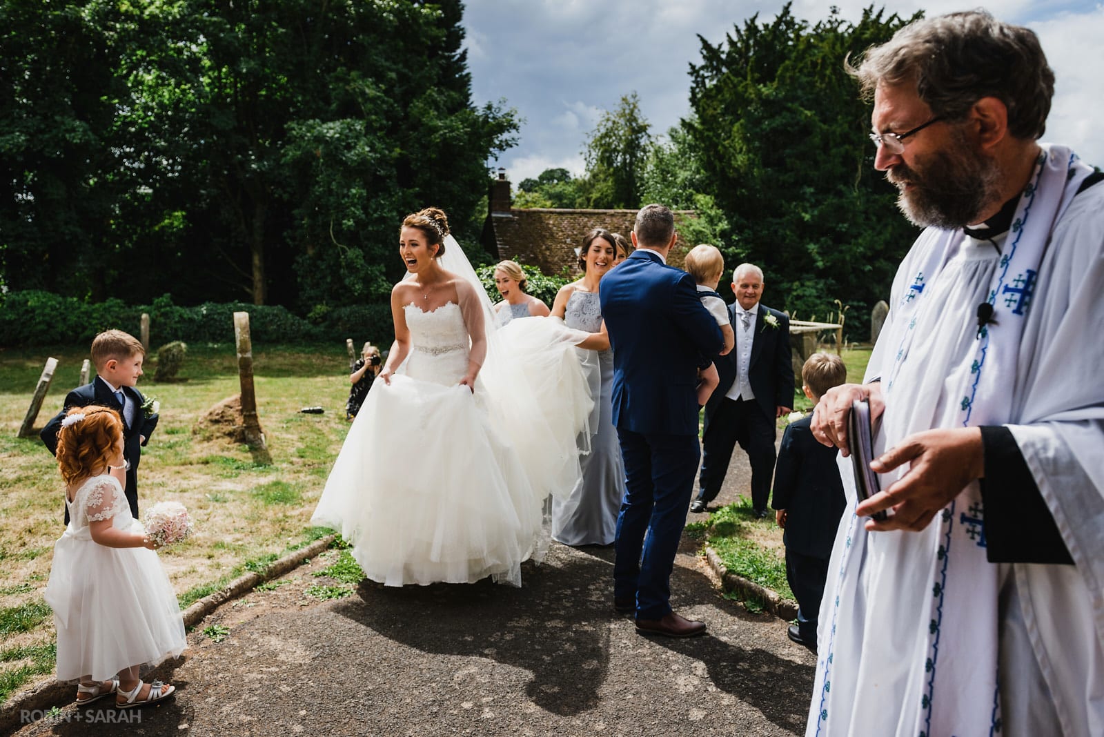 Bride greets flowergirl and pageboys outside church as vicar looks on