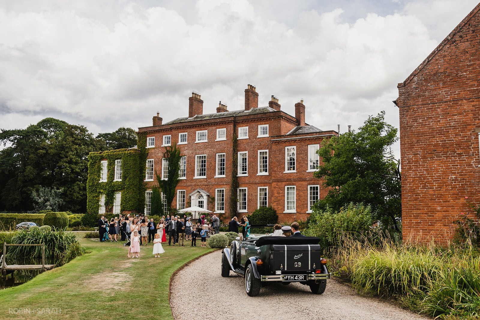 Bride and groom in open-top car arrive at Delbury Hall for wedding reception