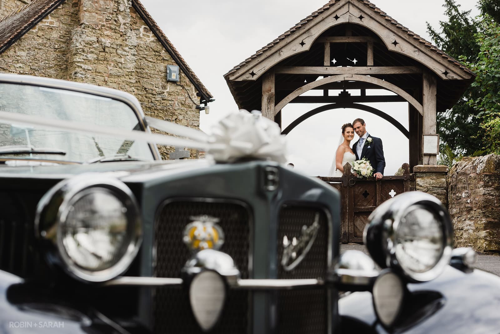 Bride and groom under lychgate at church with wedding car in foreground
