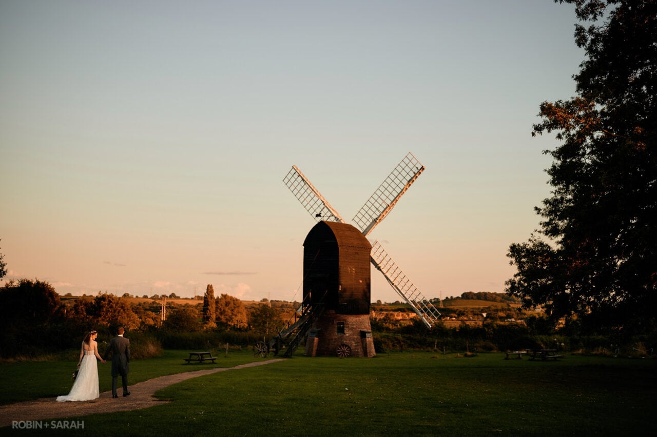 Bride and groom walk hand in hand towards windmill at Avoncroft Museum in a beautiful summer evening