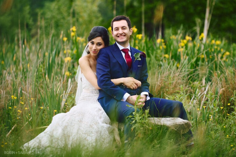 Bride and groom on wooden bench surrounded by reeds and grass in beautiful park