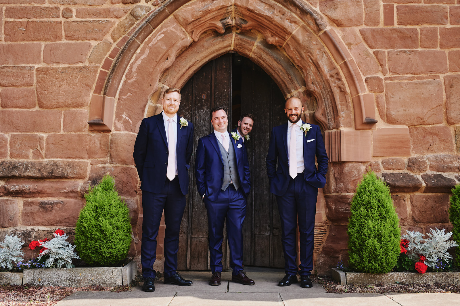 Groom and best men pose for photo outside Peter's church Dunchurch as friend pokes his head around the door