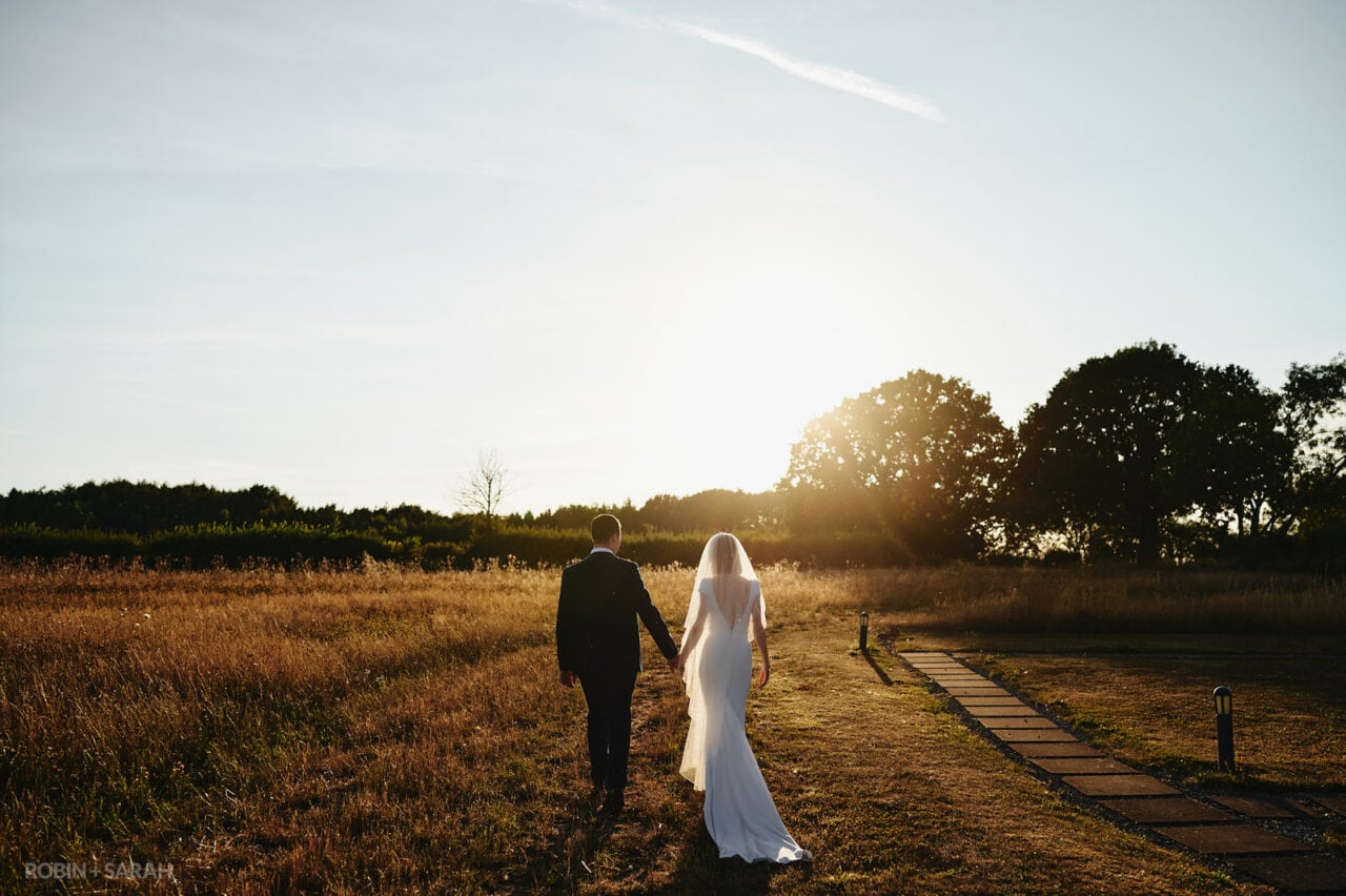 Bride and groom walk through fields towards setting sun as the light catches the bride's veil